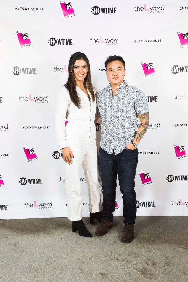 Arienne Mandi (in a white jumpsuit and black ankle boots) and Leo Sheng (in dark jeans and a black-and-white patterned button-up shirt) posing in front of a promo poster for "The L Word: Generation Q."