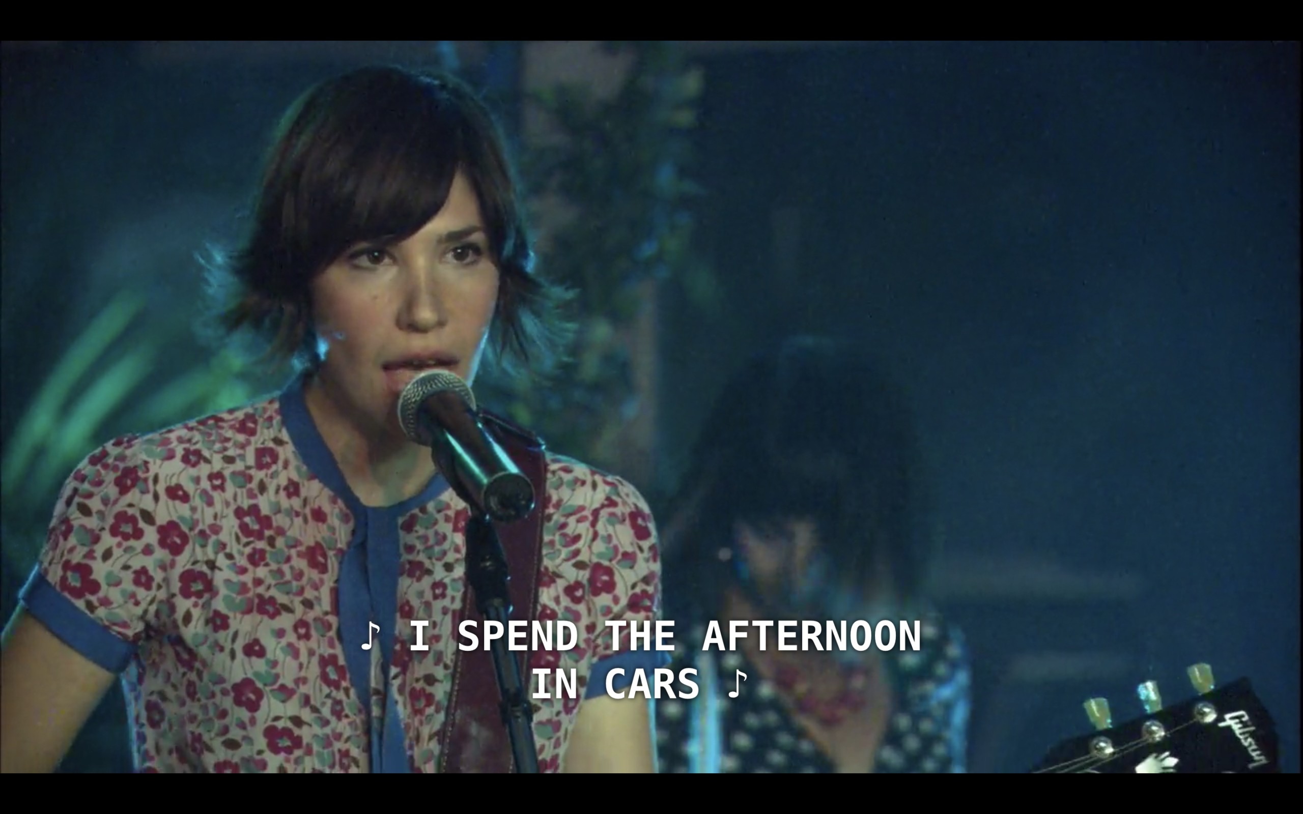 Carrie Brownstein (playing herself in Sleater-Kinney) stands at the microphone on stage performing a concert. She is holding her guitar and has brown short hair that flips out at the ends, and she is wearing a short-sleeve floral shirt. She sings, "I spend the afternoon in cars"