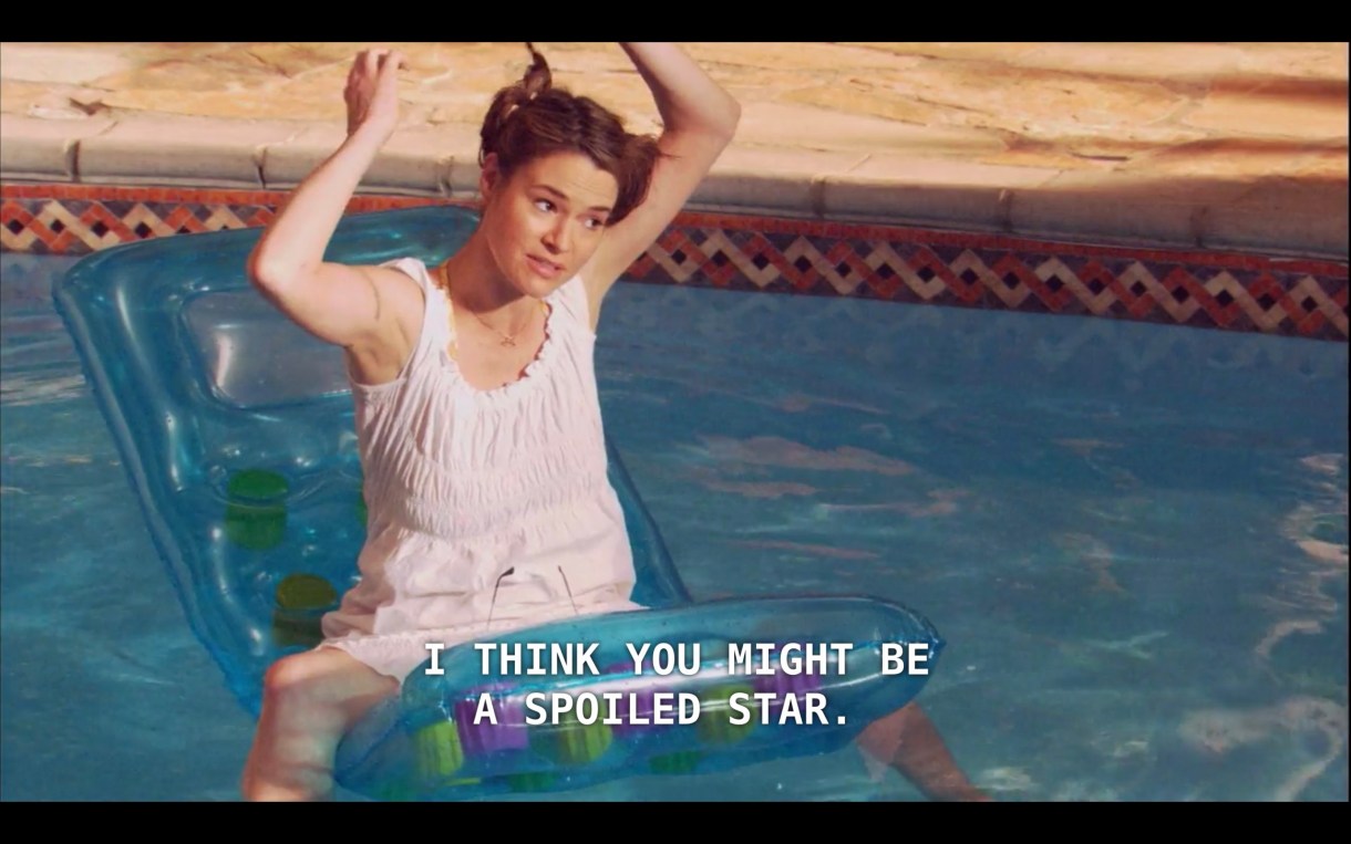 Alice (wearing a white sun dress) is straddling a pool raft in Bette and Tina's pool. Her arms are raised, and she says, "I think you might be a spoiled star."