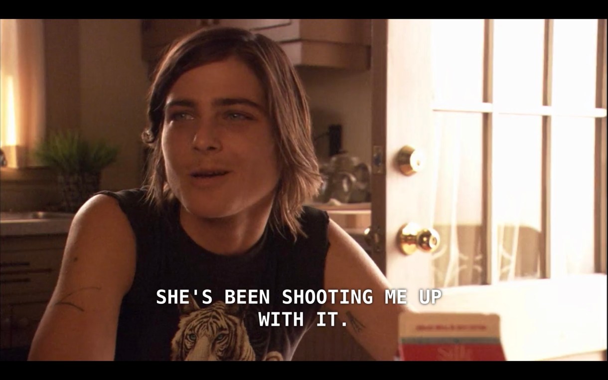 Max is wearing a black tank top with a lion on it and is sitting in the kitchen. He says, "She's been shooting me up with it."