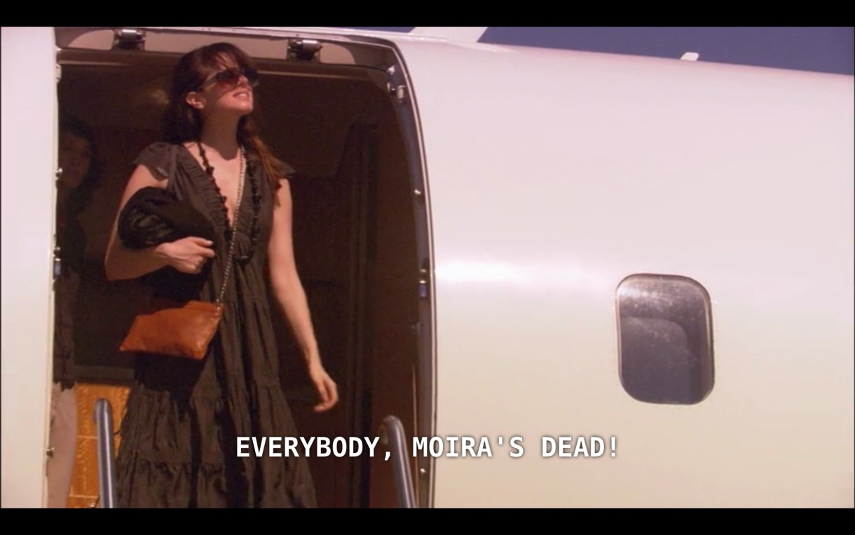 Jenny (wearing a dark brown dress and sunglasses) steps off a private plane. She says, "Everybody, Moira's dead!"