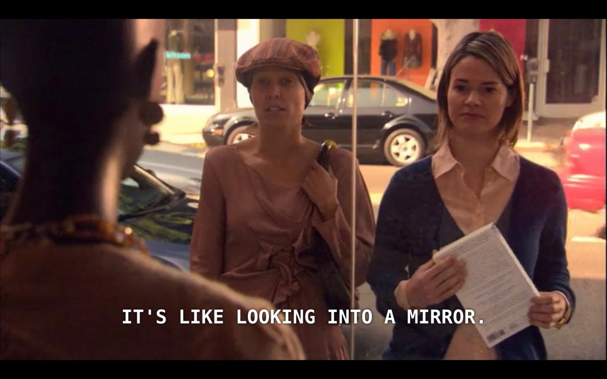 Dana and Alice are looking in a storefront window at a bald mannequin (whose back is to the camera). Dana says, "It's like looking into a mirror."