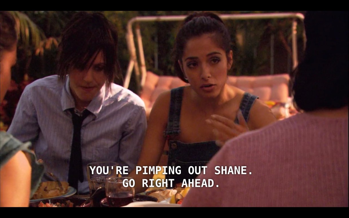 Shane (wearing a blue button-up and a skinny black tie) sits next to Carmen (wearing jean overalls) at a picnic table where they're having dinner with Carmen's family. Carmen says to her mom, "You're pimping out Shane. Go right ahead."