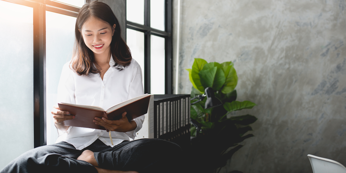 a woman reading in a bright window with a plant nearby