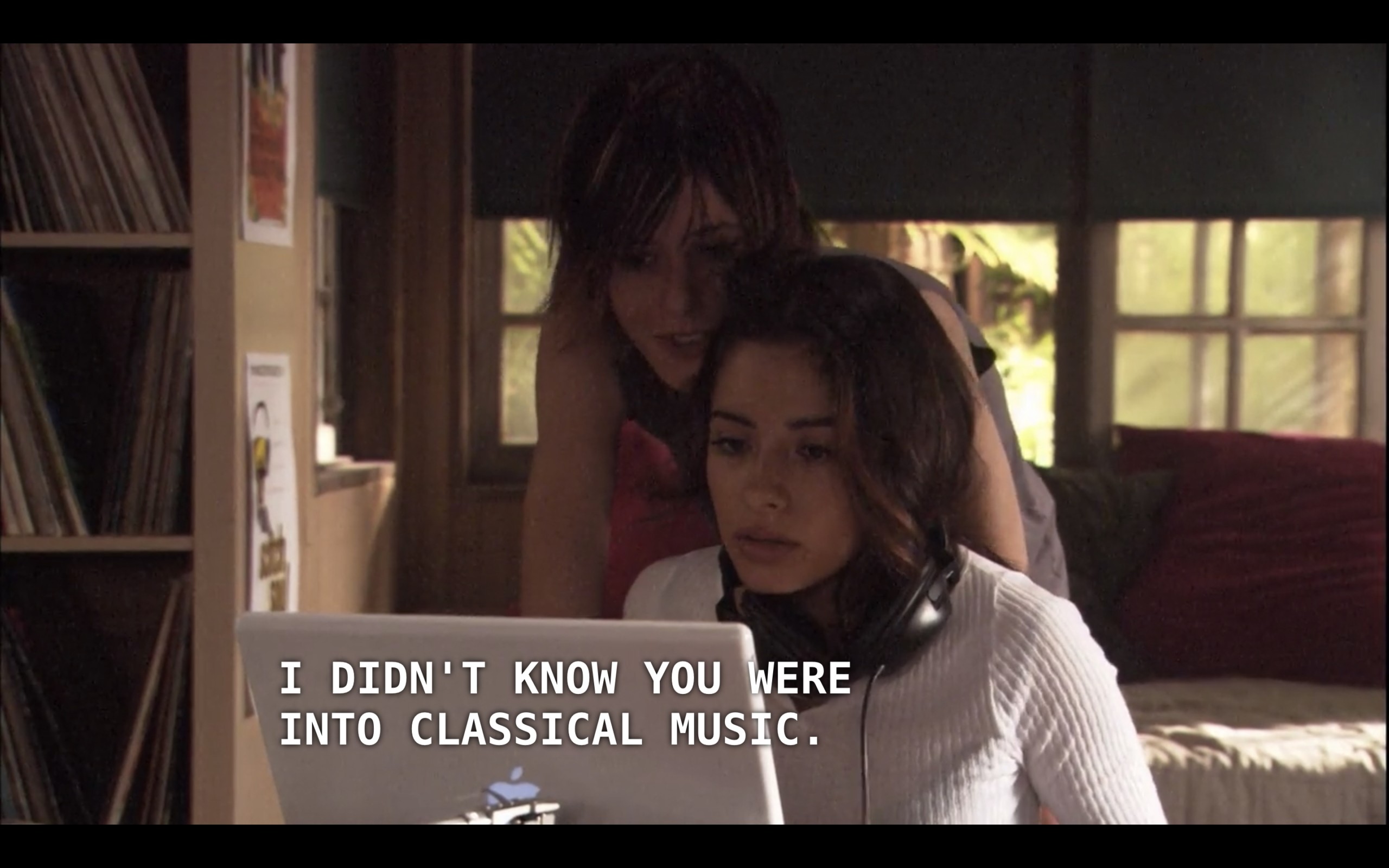 Carmen is sitting with her computer at a table in Shane and Jenny's house. She is wearing a white sweater and her headphones are hanging around her neck. Shane is looking over her shoulder and says, "I didn't know you were into classical music."