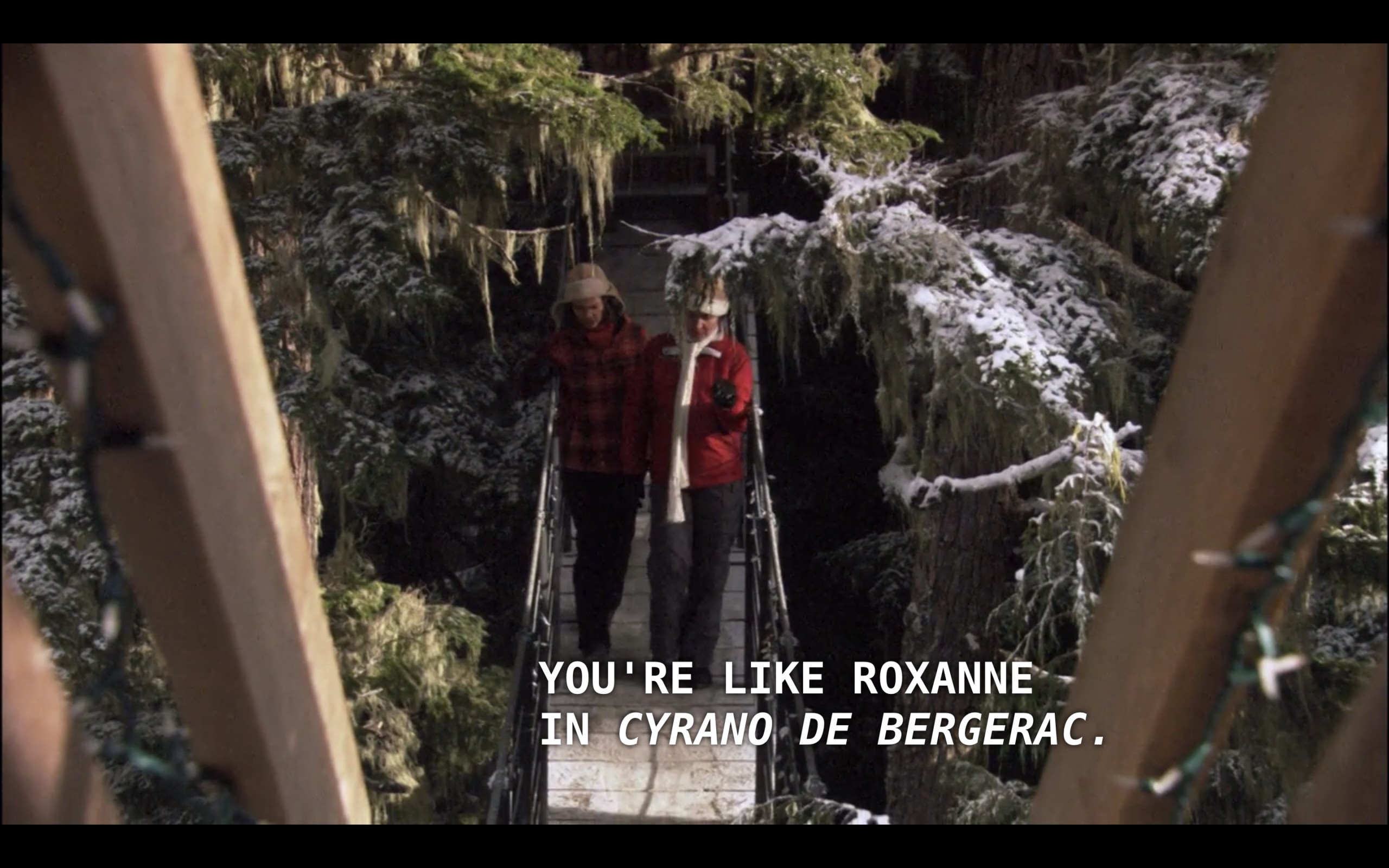 Alice (wearing a red plaid winter jacket and a brown ear-flap hat) walks next to Marilyn (a woman Alice just met, wearing a red winter jacket and white scarf) walk across a wooden bridge amongst snow-covered evergreen trees. Marilyn says, "You're like Roxanne in 'Cyrano de Bergerac.'"