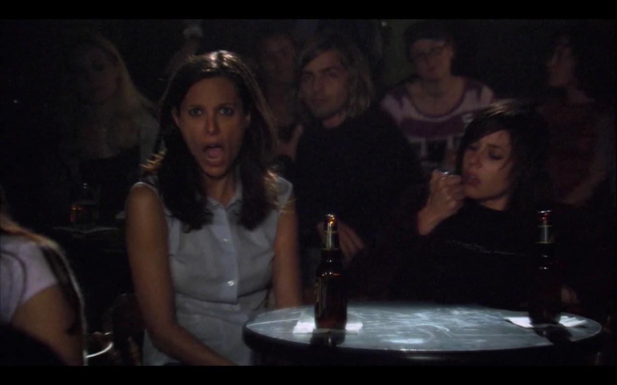 Dana and Shane sit at a table at a crowded Tegan and Sara concert. Dana's jaw has dropped, while Shane inspects her fingernail.