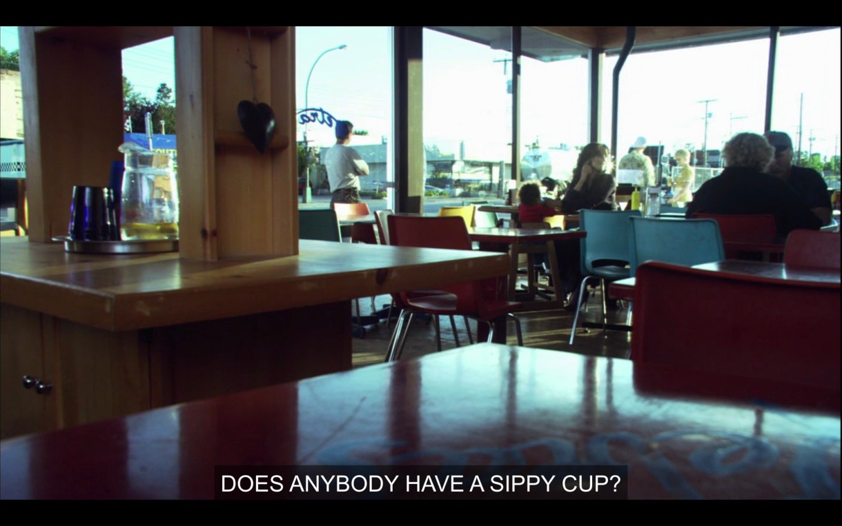 Bette Porter in a large empty cafe with her baby, on her cell phone.