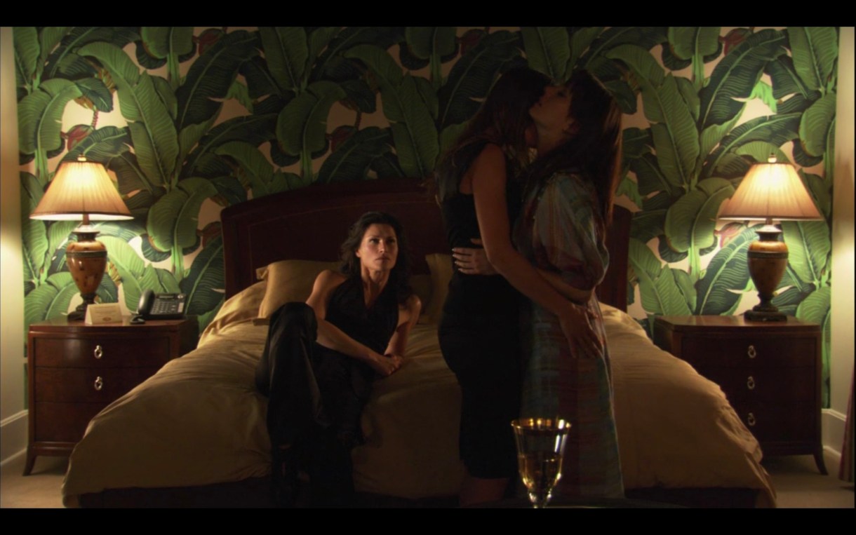 In a hotel room with wallpaper that looks like a forest. Marina is lying on the bed. Dim lighting. Claude and Jenny are standing up, making out. 