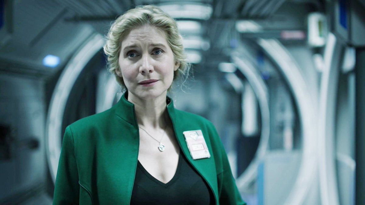 Image: a middle-aged white woman in a black v-neck shirt and green blazer stands in what appears to be the hallway of a spaceship. She looks concerned.