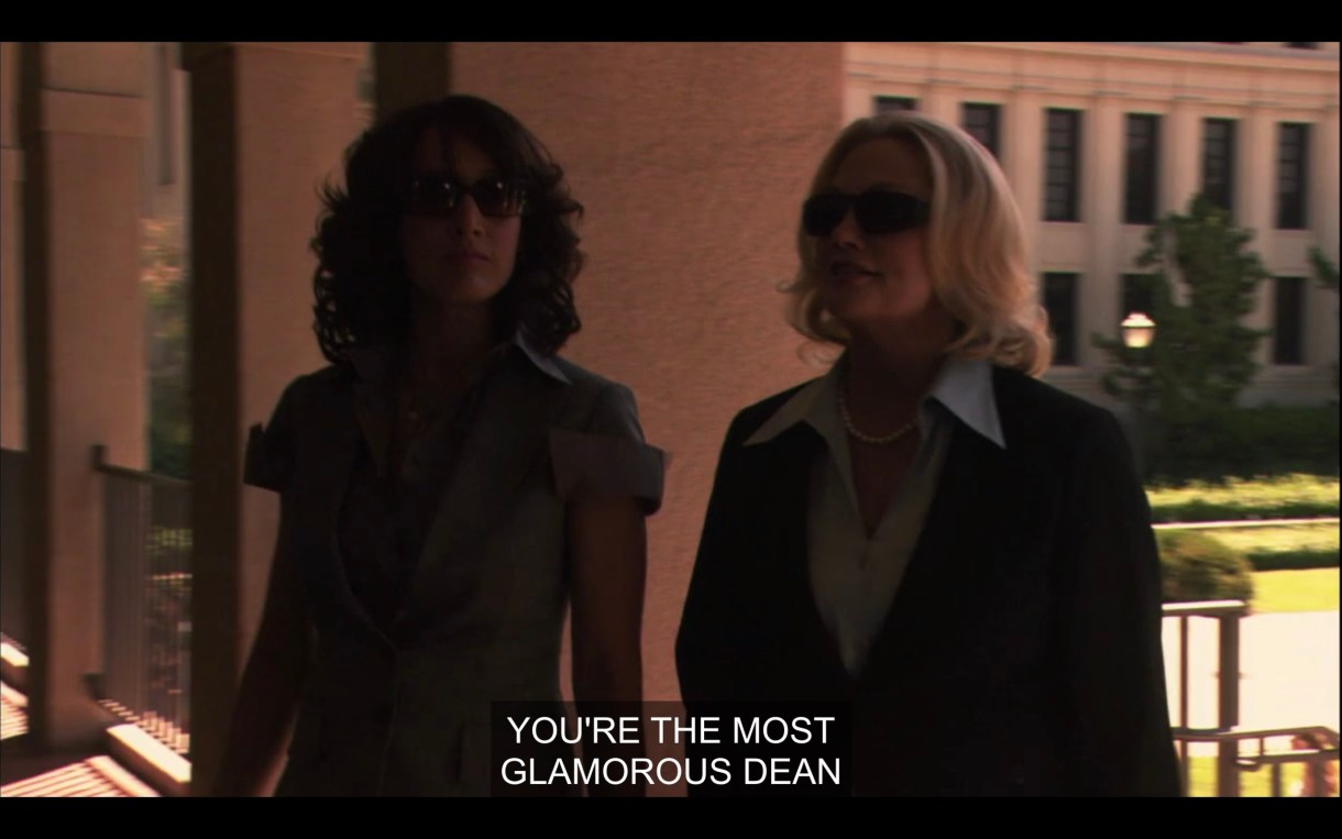 Bette and Phyllis, both wearing dark sunglasses, are walking into a building from outside. Phyllis says, "You're the most glamorous dean."
