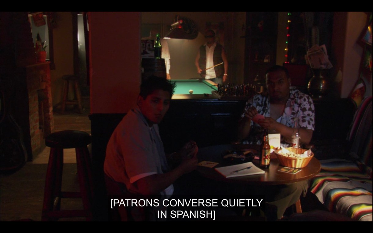 A dimly lit bar with a pool table in the background and two guys sitting at a table. Subtitles read, "[Patrons converse quietly in Spanish.]"