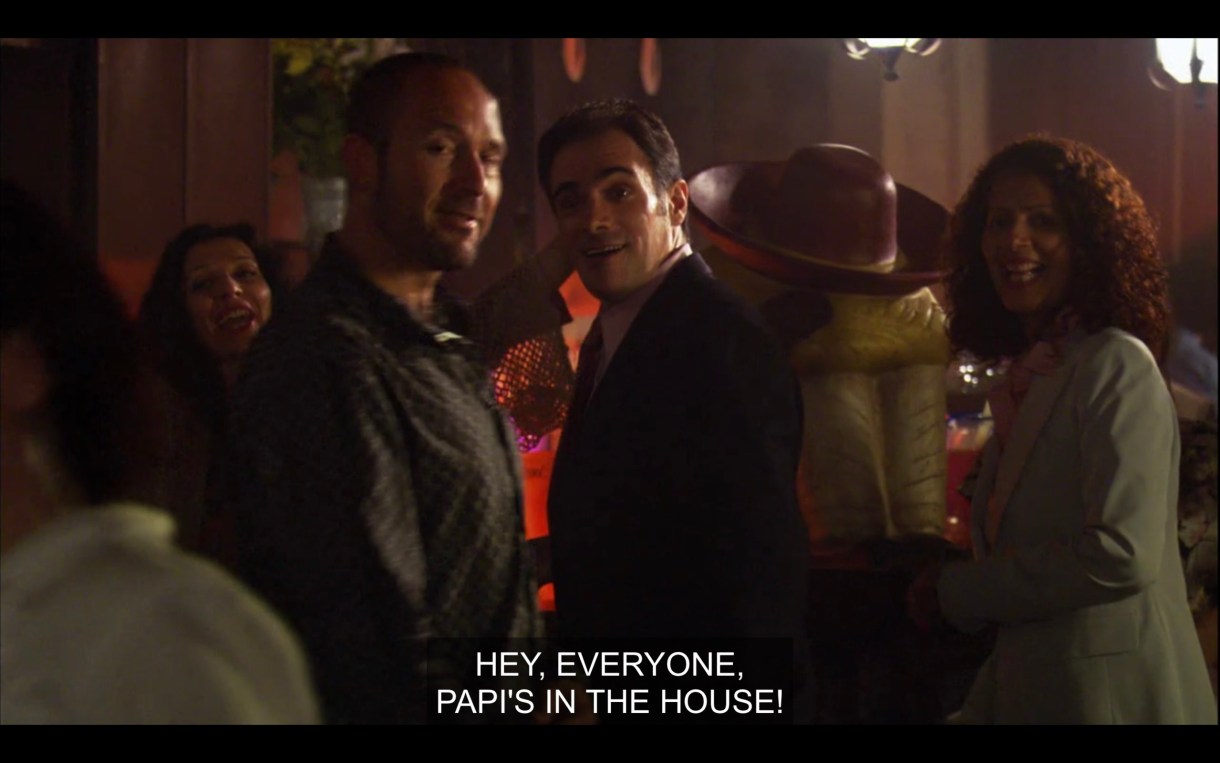 A crowd of unknown people at a bar. One of them shouts, "Hey, everyone, Papi's in the house!"