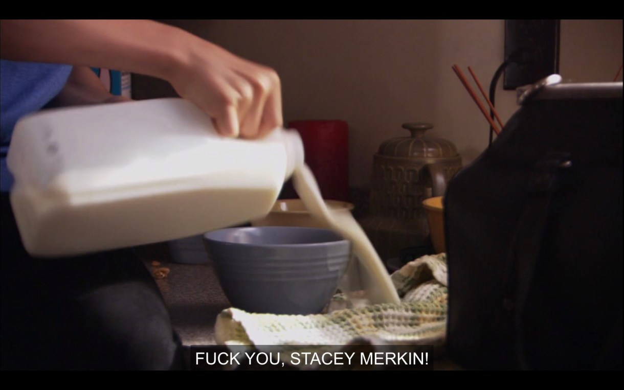 A hand messily pours milk from a jug into a blue cereal bowl. Jenny yells off camera, "Fuck you, Stacy Merkin!"