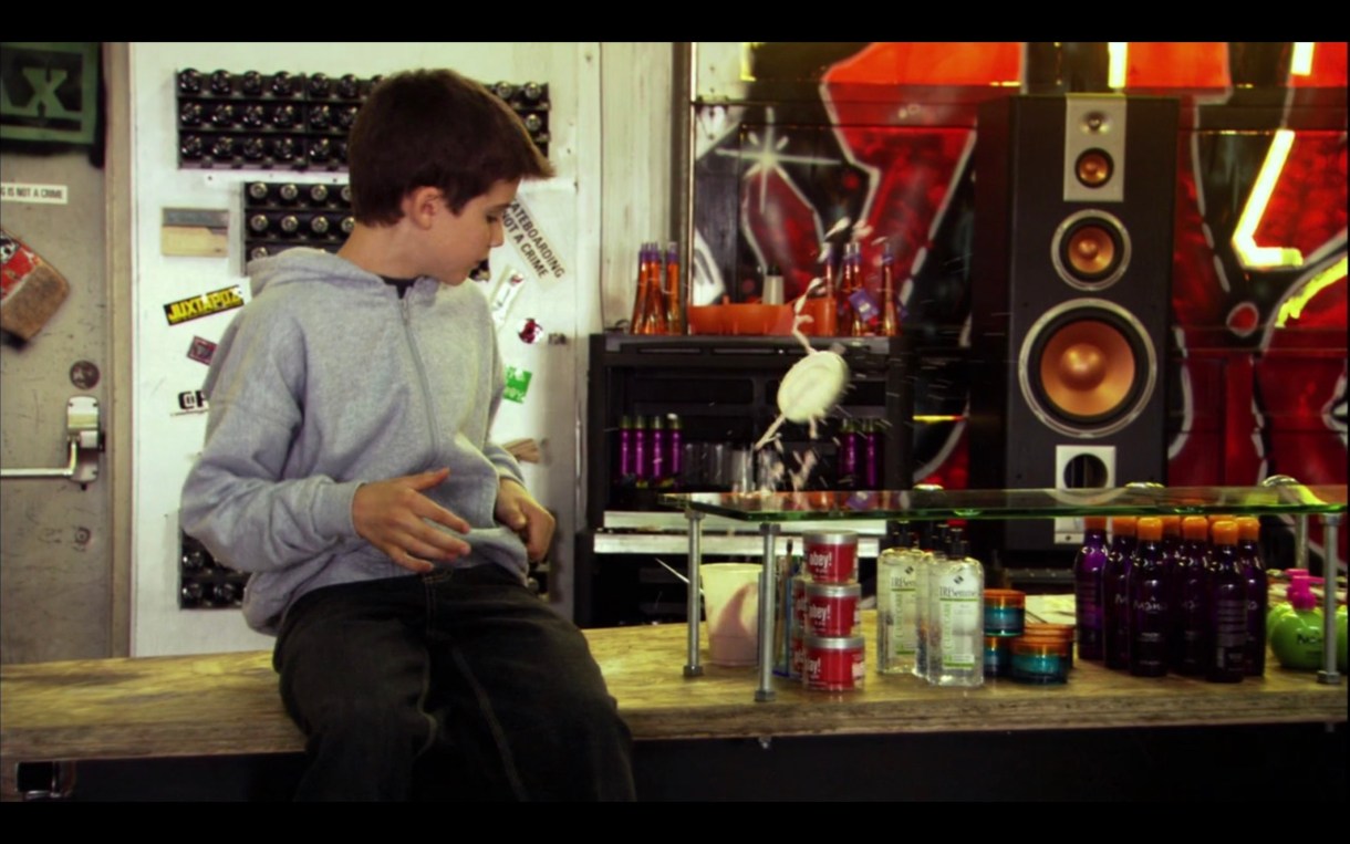 Shay sits on the counter at the skate shop, wearing a grey hoodie. He's looking at the haircare products on display.