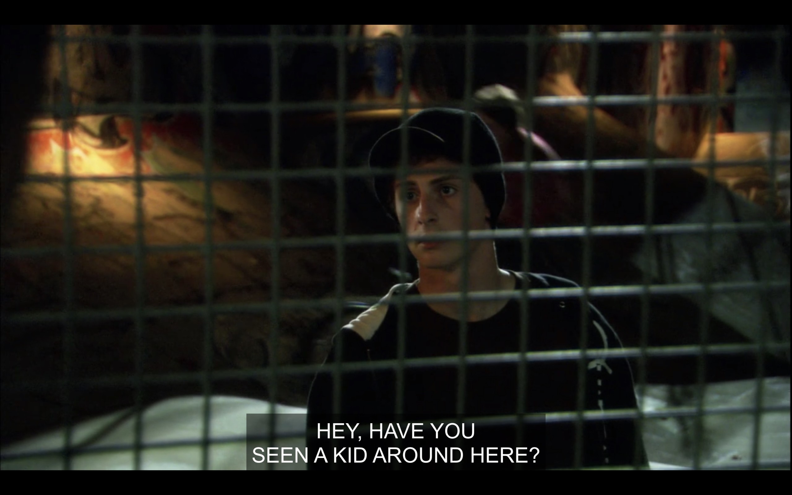 A skater due in a black hoodie and hat, standing behind a fence near a skateboard ramp. Max, off camera, asks him, "Hey, have you seen a kid around here?"