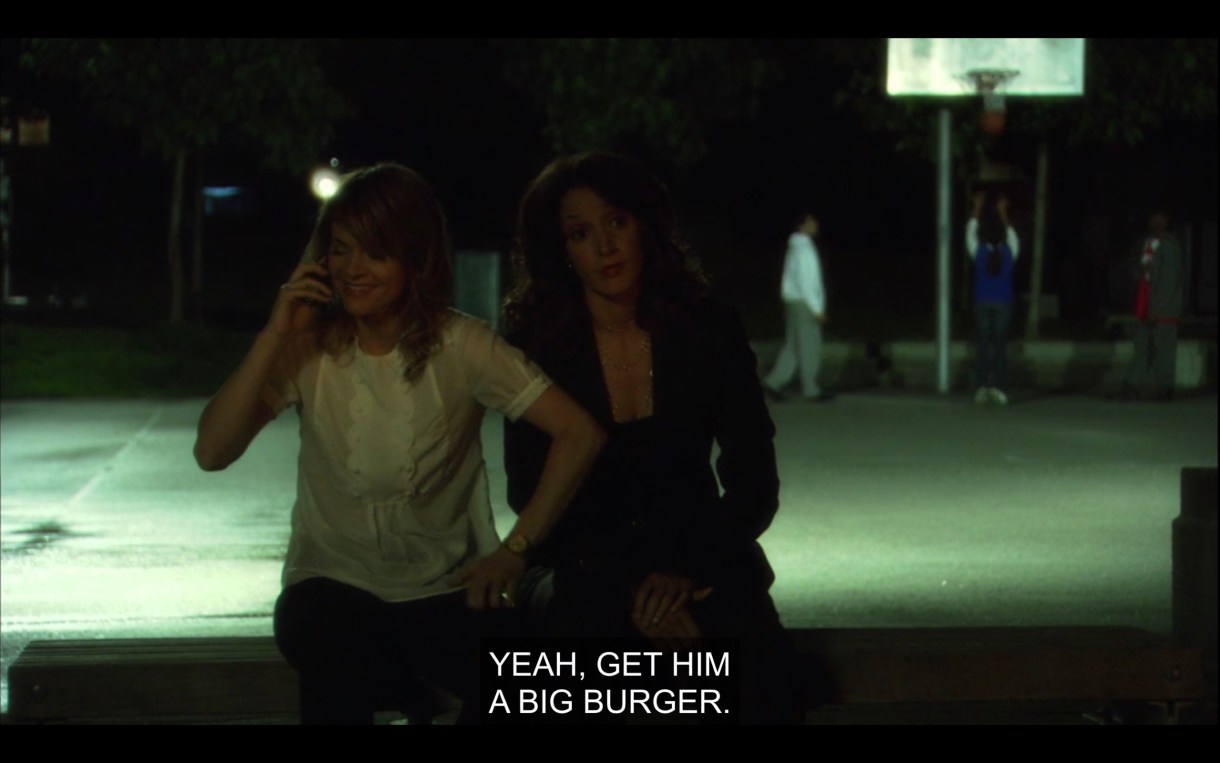 Alice and Bette sit on a bench near a basketball court. Alice is talking to Shane on the phone. Alice says, "Yeah, get him a big burger."
