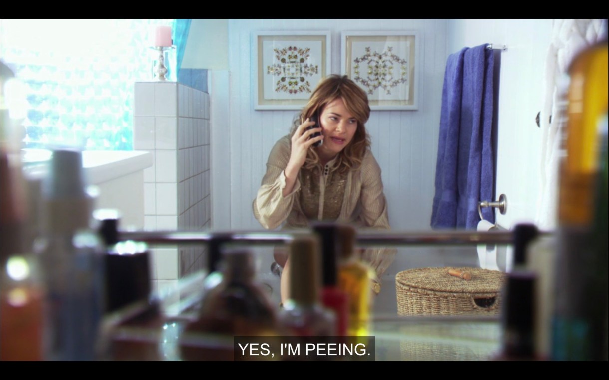 Alice is wearing a brown top and sitting on the toilet, talking on her cell phone. She says, "Yes, I'm peeing."