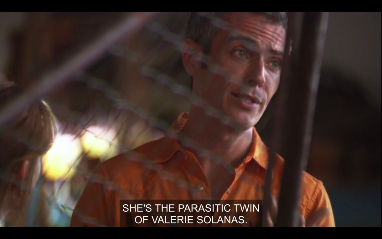 Tom wearing an orange button up. He says to Bette, "She's the parasitic twin of Valerie Solanas."