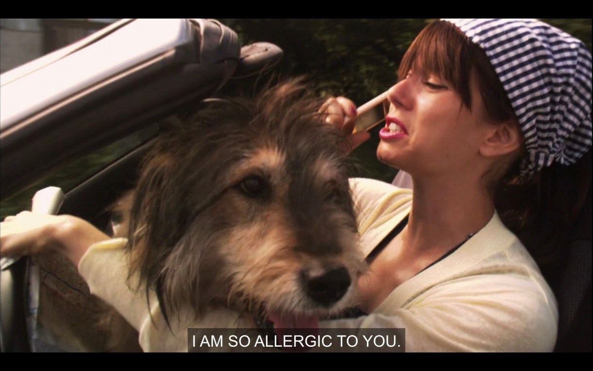 Jenny (wearing a yellow cardigan and a gingham bandana on her head) is driving a convertible with Sounder sitting in her lap. She says, "I am so allergic to you."