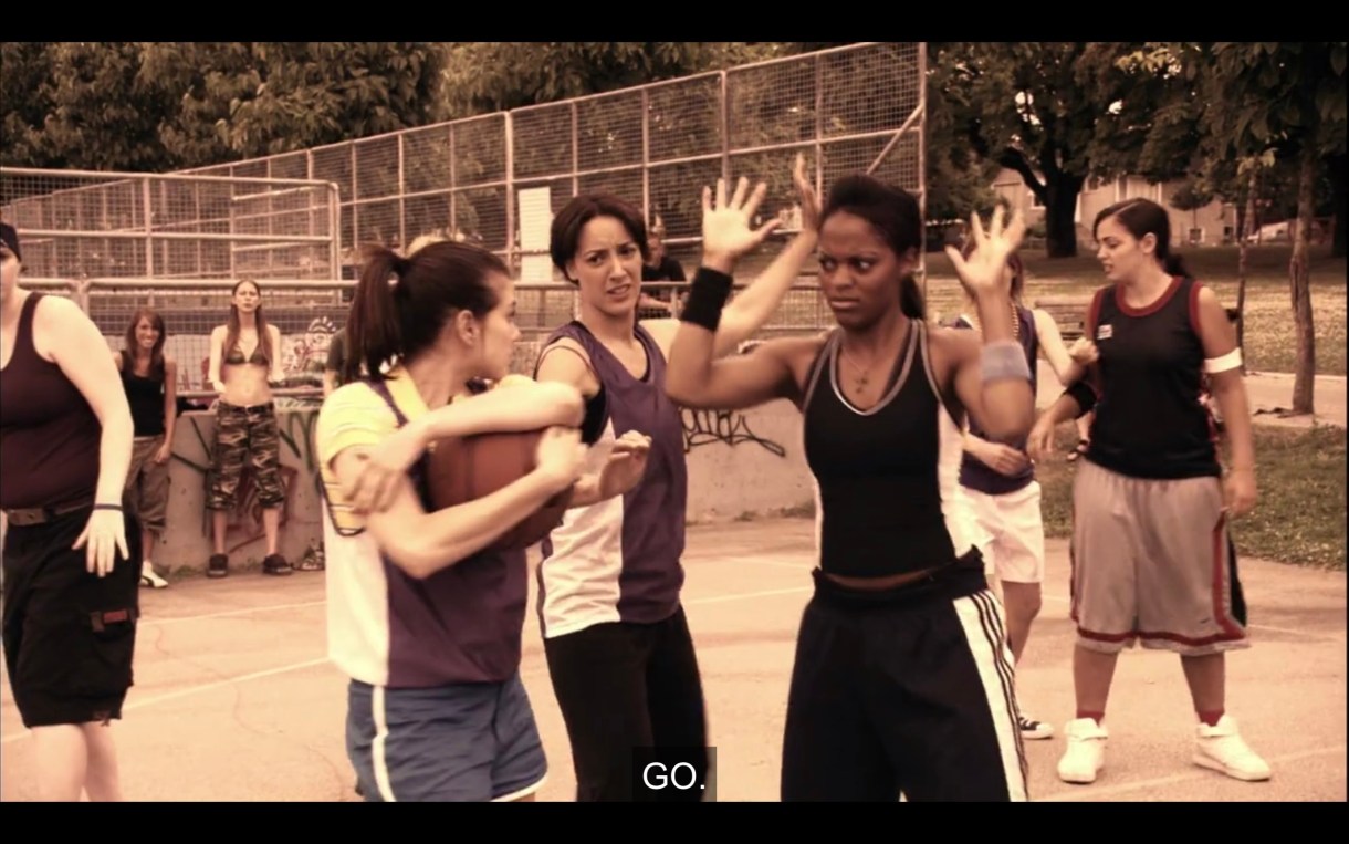 Jenny is hugging the ball close to her chest on the basketball court. One of her opponents has her hands raised in the air.