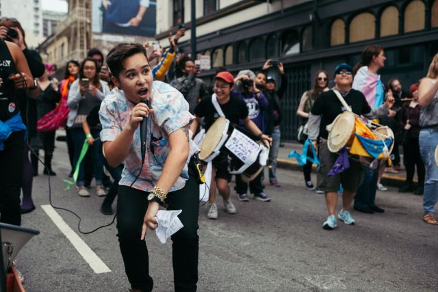 Sammie bends low to the ground to speak into a mic at a march while people behind them play drums