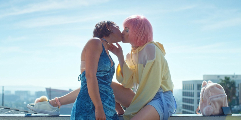 A girl with pink hair and a yellow sweatshirt kisses another girl in a blue dress with a cityscape behind them.