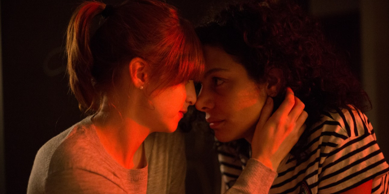 Two women lean their heads against each other in red lighting.