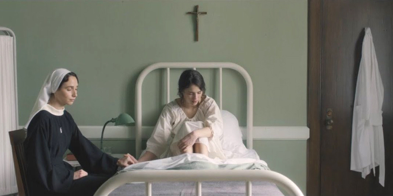 A young woman in white sitting in bed below a cross holds hands with a nun.