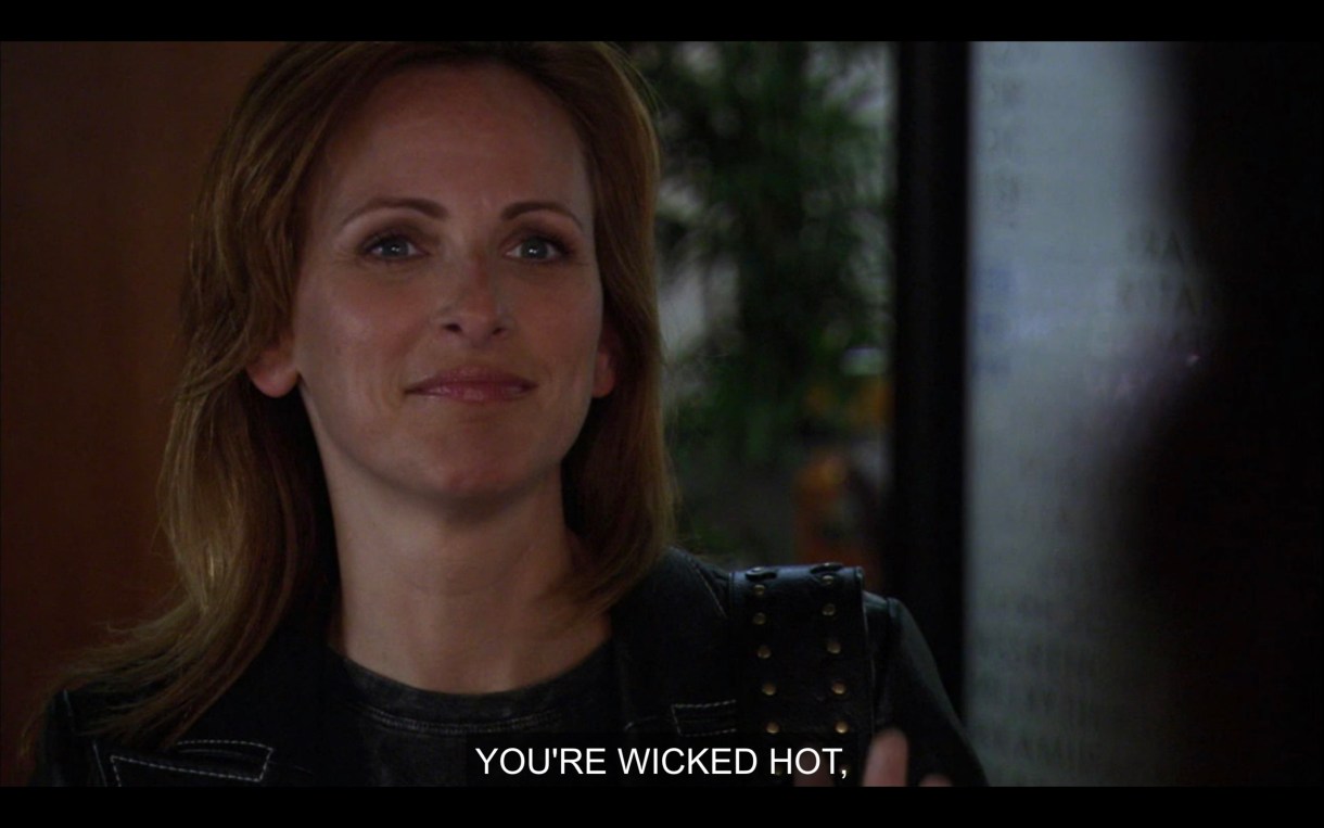 Jodi, in a black blazer, stands in Bette's office. She's smiling with her mouth closed. "You're wicked hot."