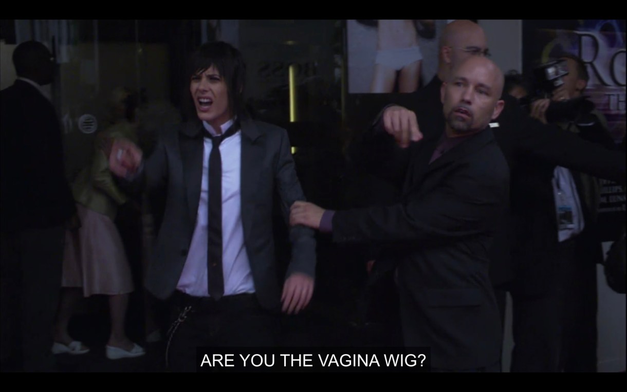 Shane (wearing a black skinny tie, white shirt, and black blazer) is having her arm held back by a security guard in all black. She shouts, "Are you the vagina wig?"