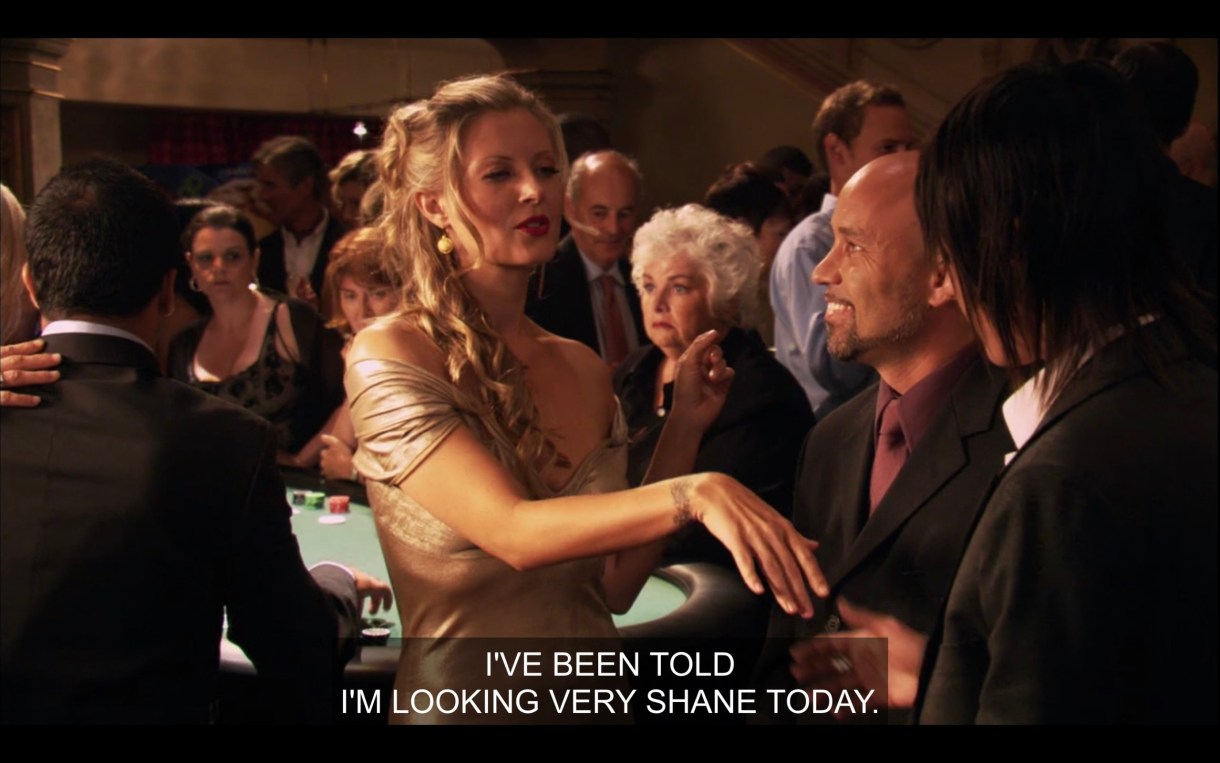 Shane shakes the hand of a woman with long blonde hair and wearing a ball gown, standing in front of a poker table. She says to Shane, "I've been told I'm looking very Shane today."