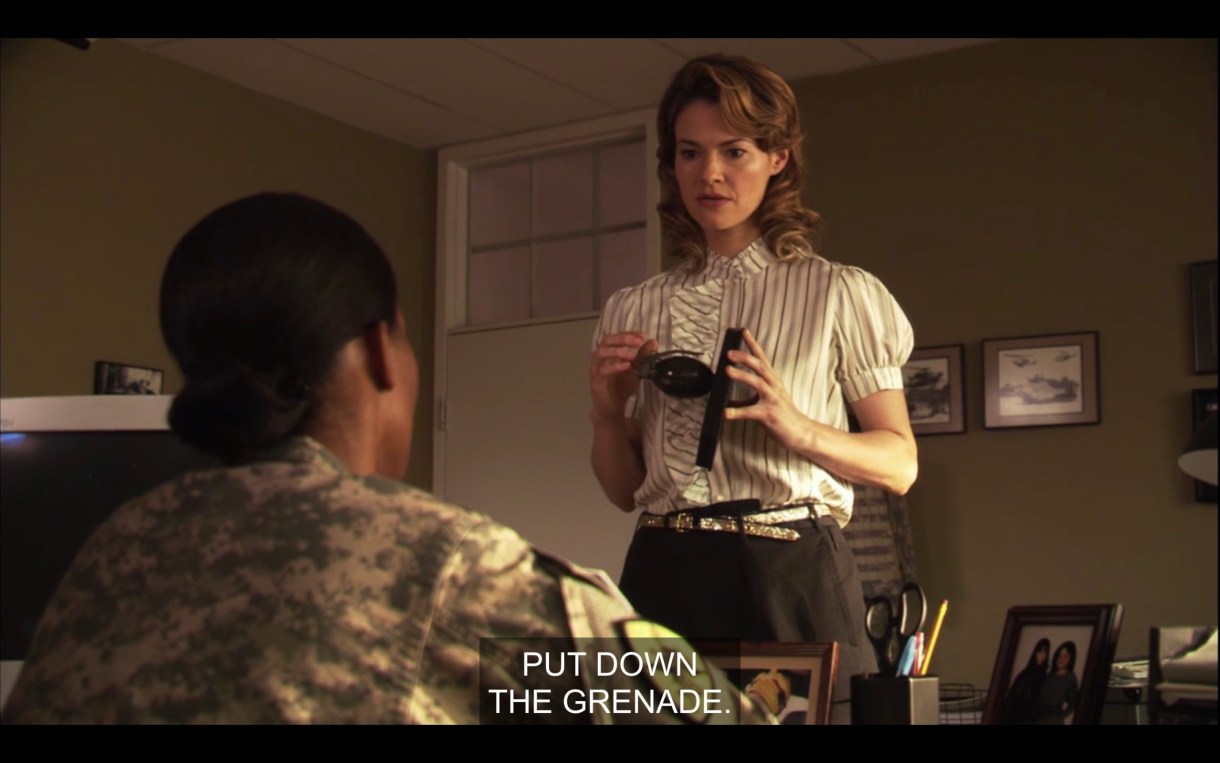 In Tasha's office, Alice is standing in front of Tasha'a desk holding a grenade. Alice's hair is very 50s. The caption reads "put down the grenade."