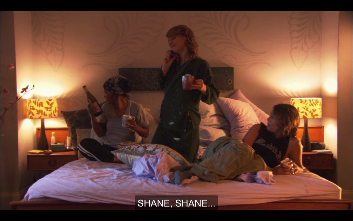 Tasha, Alice, and Papi in Alice's bed. Tasha is holding a bottle of wine. Tasha and Alice are both holding coffee cups. Alice is talking on the phone. She says, "Shane, Shane..."