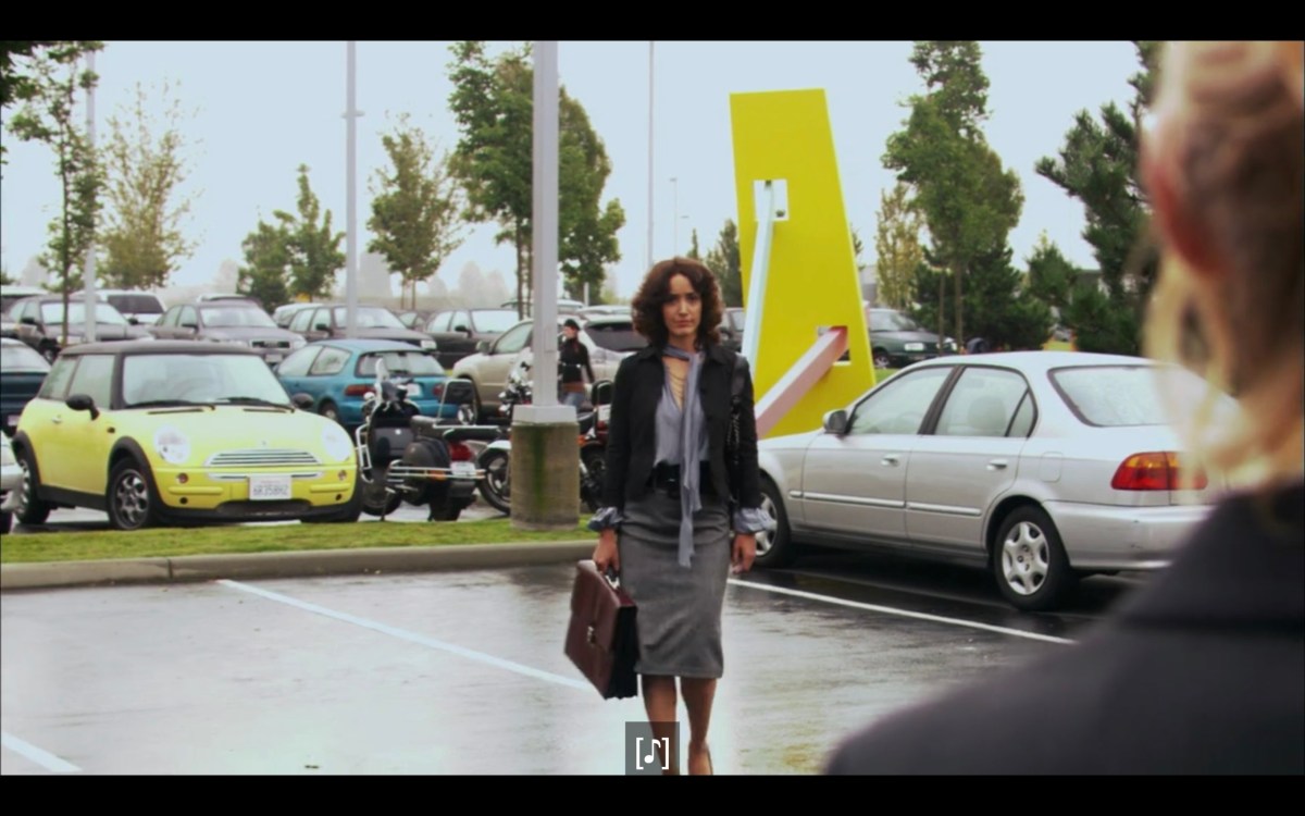 Bette (in a grey skirt, black jacket, carrying a brown brief case) walking through a parking lot towards Tina (who is barely in frame). 