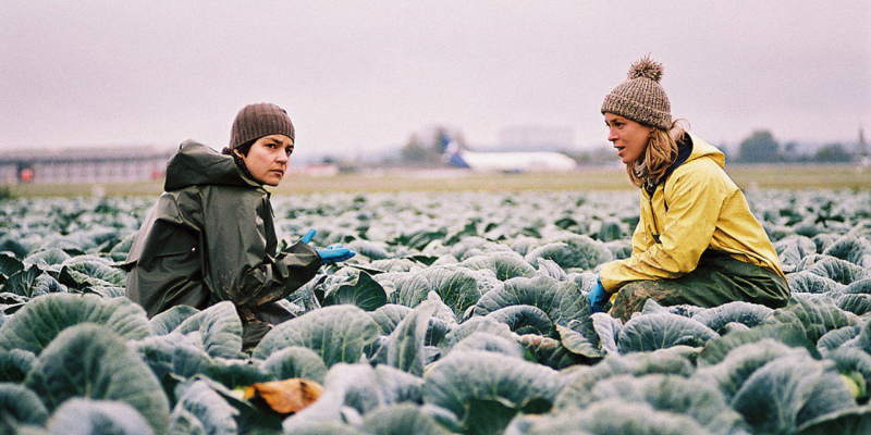 Two women in beanies stand among leafy crops.