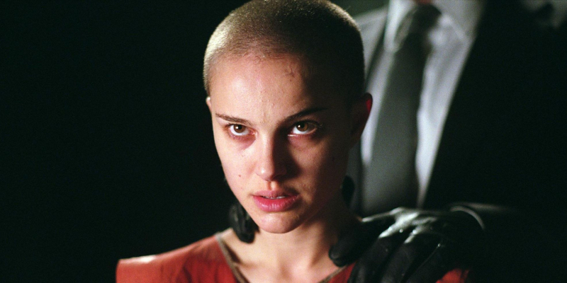 Natalie Portman looks up with a shaved head and gloved hands on her shoulder.