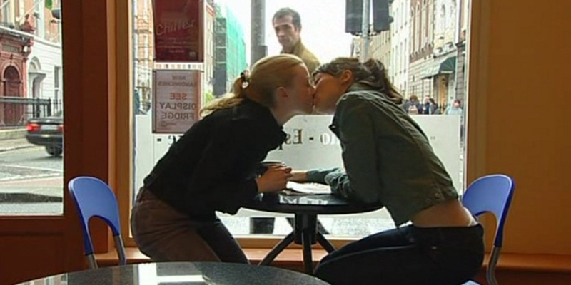 Two women kiss in a cafe while a man looks at them through the window.