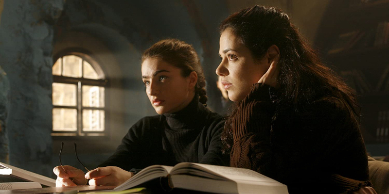 Two girls sit at a table with a pile of open books.