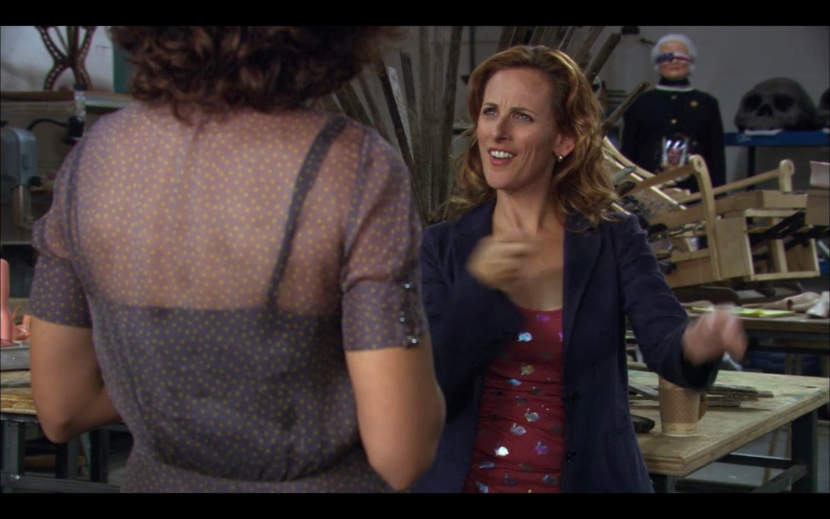 Bette (wearing a gray blouse, her back to the camera) is talking with Jodi (wearing a pink top under a blue cardigan) in Jodi's studio. Behind Jodi is her sculpture of Barbara Bush, who is blindfolded with an American flag.