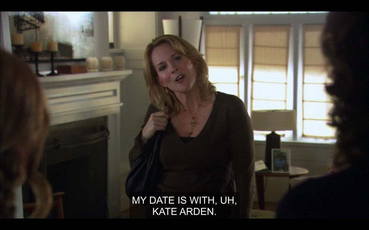 Tina in her living room, wearing a brown top and a purse over her shoulder. Her head is tilted to her right, and is facing Jodi and Bette (who are off camera). Tina says, "My date is with, uh, Kate Arden."