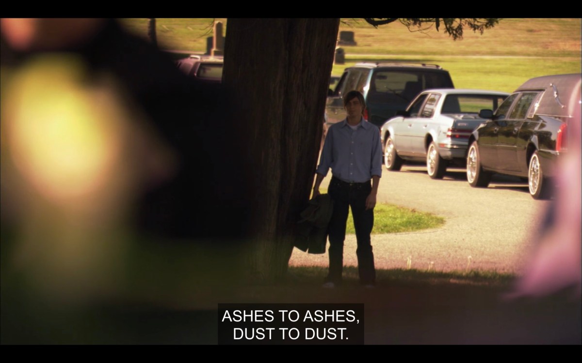 Max (wearing a blue dress shirt) is walking along a dirt path. Subtitles read, "Ashes to ashes, dust to dust."