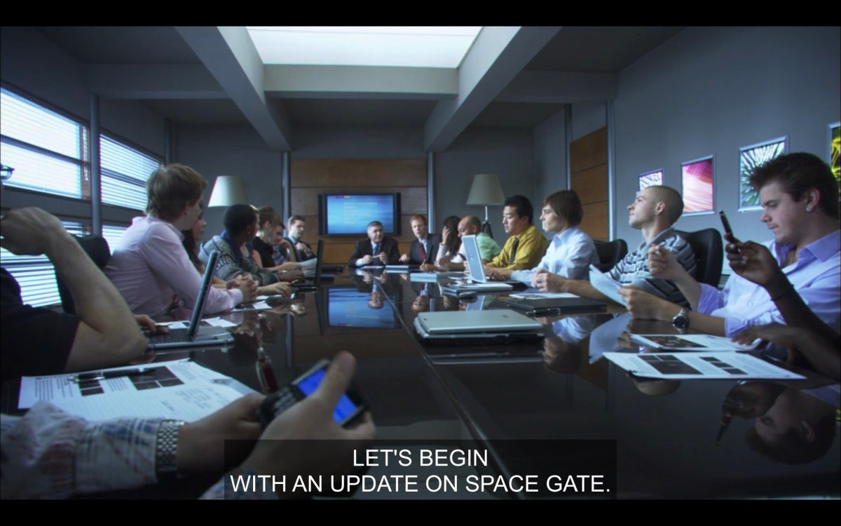 A big long conference table, with Max surrounded by a bunch of other tech dudes on their phones and laptops. His boss, at the head of the table, says, "Let's begin with an update on space gate."