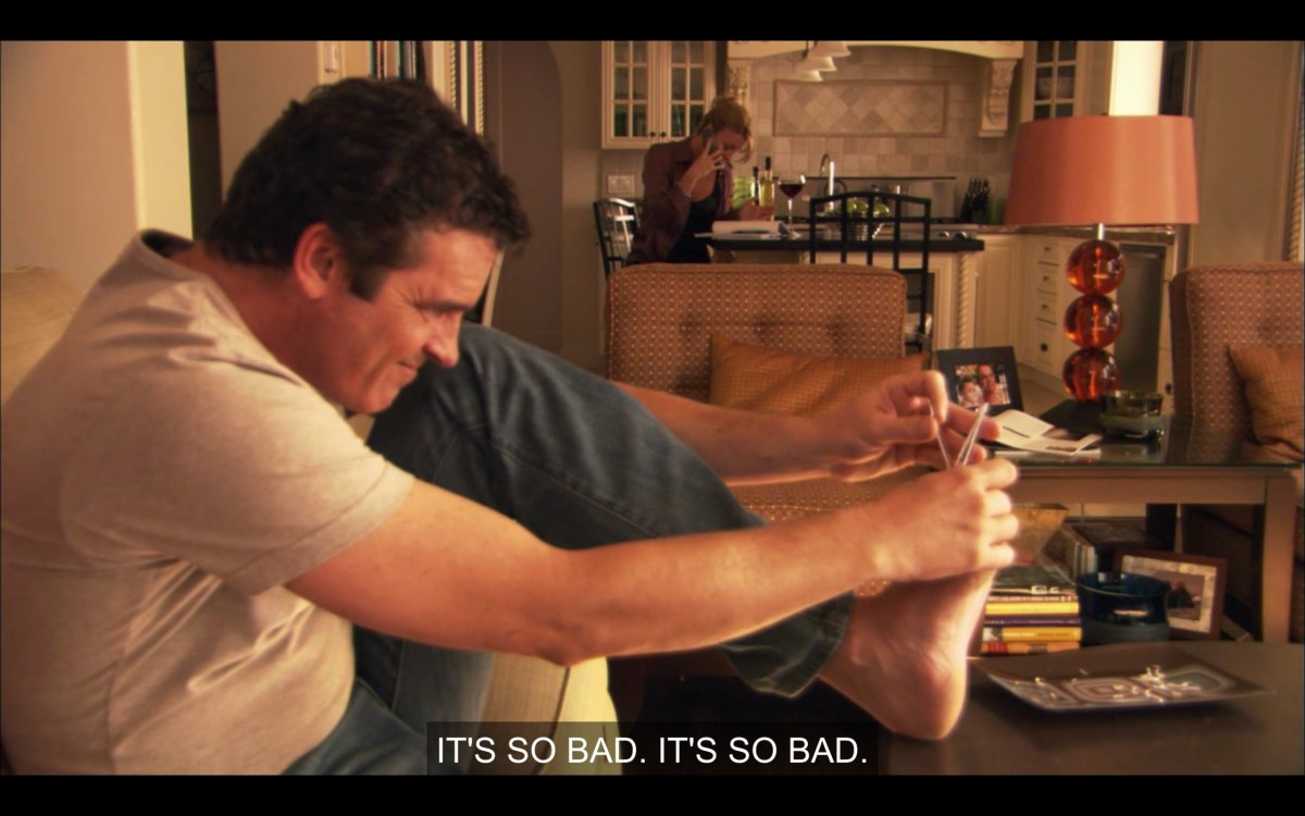 In the foreground, Henry is clipping his toenails on the coffee table. Behind him, Tina is hunched over the counter talking on her cell phone.  She says, "It's so bad. It's so bad."