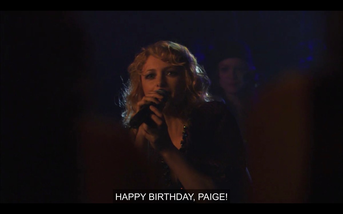 A blonde singer in a dark room holding a microphone. She says, "Happy birthday, Paige!"