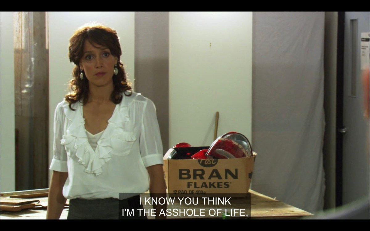 Bette in a white frilly top, standing in front of one of Jodi's work benches, saying, "I know you think I'm the asshole of life."