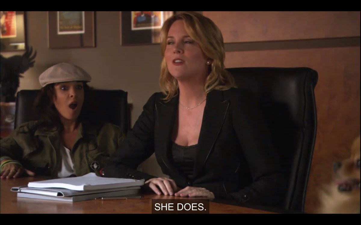 Tina (in a black suit) sits at a conference table in front of a stack of white papers. There's a brunette woman in a tan cap sitting next to her with her mouth wide open. Tina says, "She does."