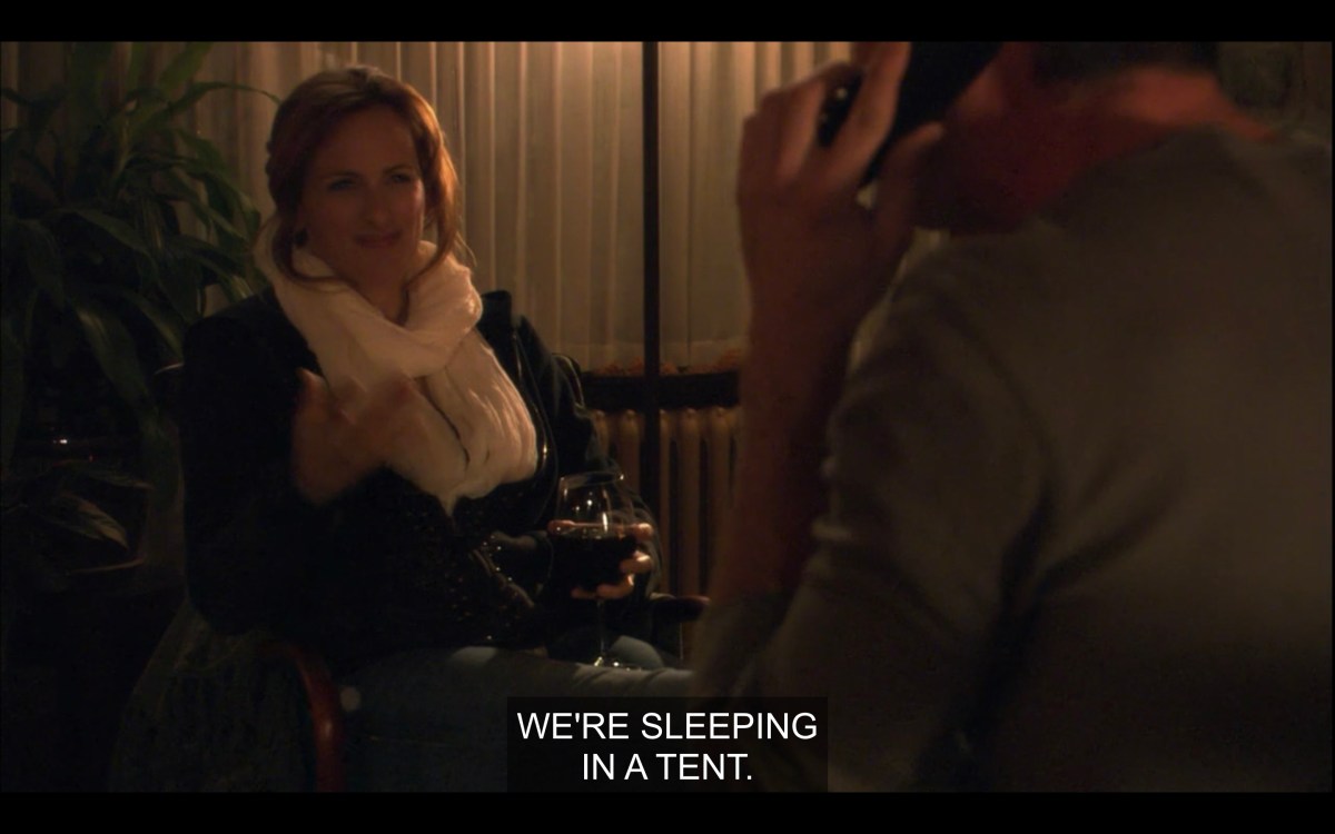 Dimly-lit shot of Jodi, holding a glass of red wine and wearing a big white scarf. Bette sits across from her, her back to the camera. Jodi says, "We're sleeping in a tent."