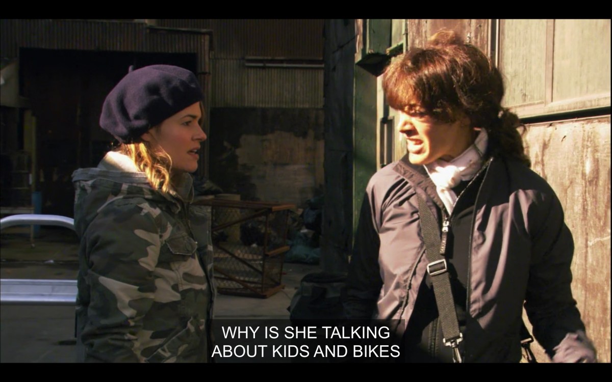 Alice (in a black beret and a camo jacket) and Bette (wearing a black jacket and a black messenger bag across her body) facing each other outside. "Why is she talking about kids and bikes?"