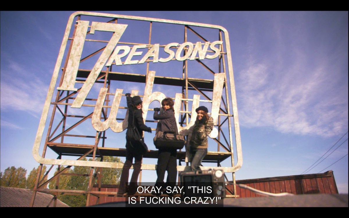 Shane, Bette, and Alice stand in front of a giant billboard that says, "17 reason why!" They're posing for a picture. Alice says, "Okay, say, 'This is fucking crazy!'"